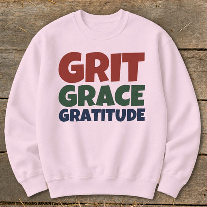 A light pink crewneck sweatshirt featuring the stacked words GRIT, GRACE, and GRATITUDE on a rustic wooden background.