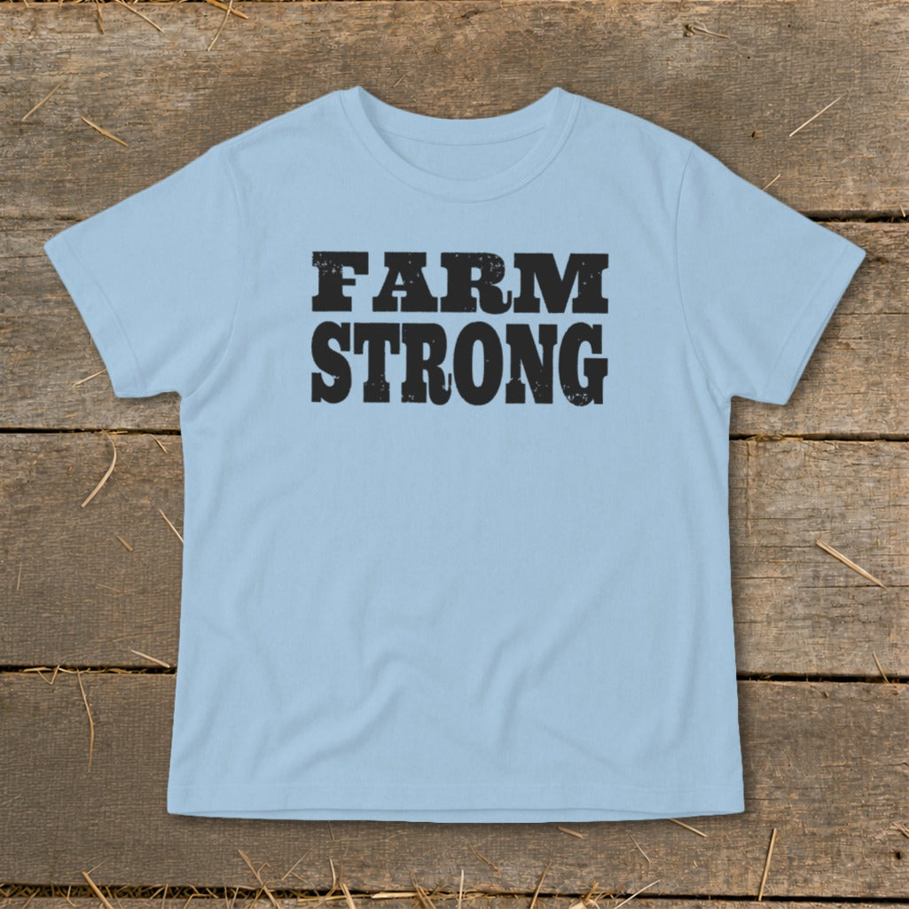 Light blue t-shirt featuring the black distressed text FARM STRONG centered on the chest, laid flat on a rustic wooden background.