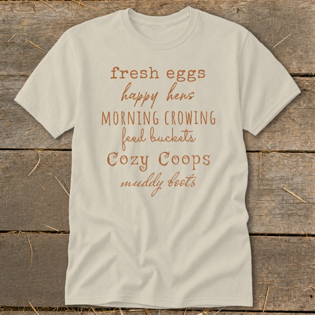 Light beige t-shirt with text: fresh eggs, happy hens, morning crowing, feed buckets, cozy coops, muddy boots.