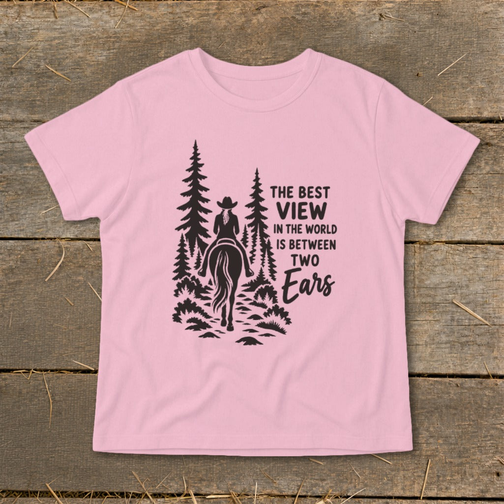 A pink t-shirt featuring a black graphic of a horseback rider and pine trees with text that reads, "The best view in the world is between two ears."
