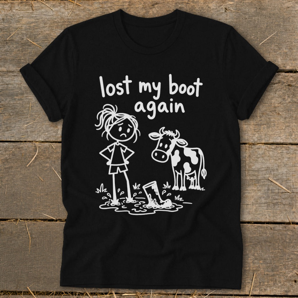 A black t-shirt printed with a white cartoon design of a girl, cow, and muddy boot alongside text reading lost my boot again.