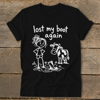 A black t-shirt printed with a white cartoon design of a girl, cow, and muddy boot alongside text reading lost my boot again.