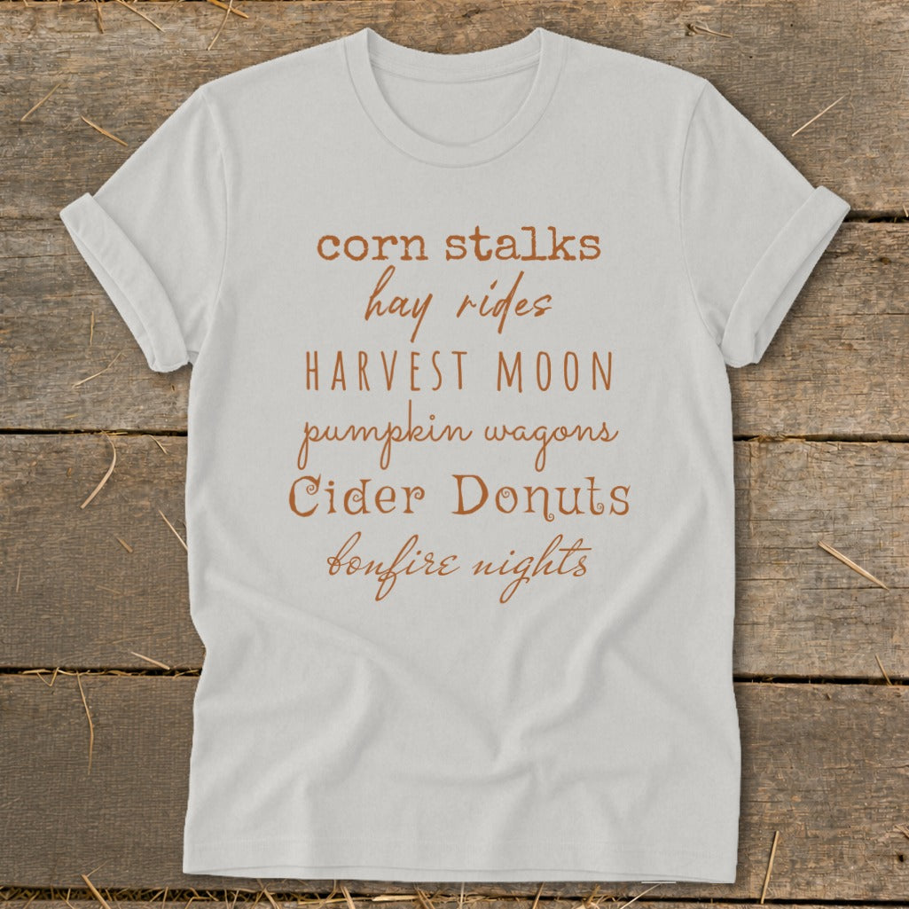 White t-shirt with "corn stalks, hay rides, Harvest Moon, pumpkin wagons, Cider Donuts, bonfire nights" text.