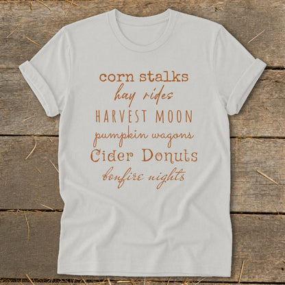 White t-shirt with "corn stalks, hay rides, Harvest Moon, pumpkin wagons, Cider Donuts, bonfire nights" text.