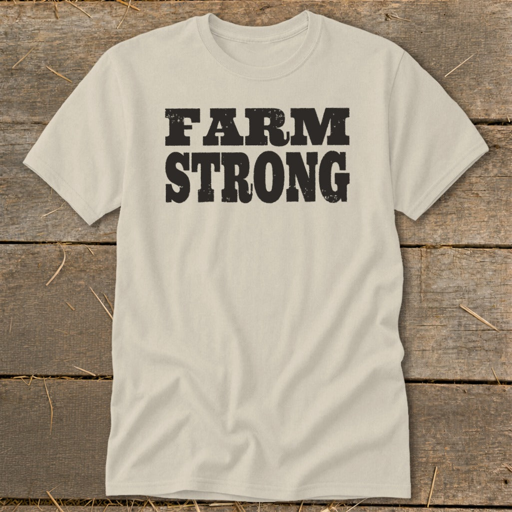 A cream-colored crewneck t-shirt with black, distressed block lettering that reads FARM STRONG centered on the chest, laid flat on a rustic wooden surface.