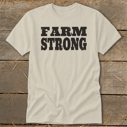 A cream-colored crewneck t-shirt with black, distressed block lettering that reads FARM STRONG centered on the chest, laid flat on a rustic wooden surface.