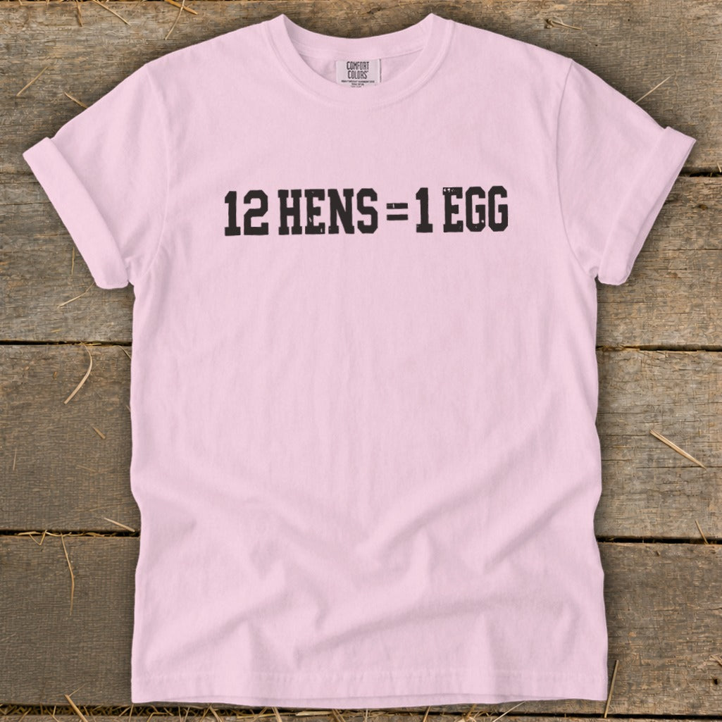 A light pink t-shirt with rolled sleeves lies flat on a wooden surface, featuring black text that reads "12 HENS = 1 EGG".