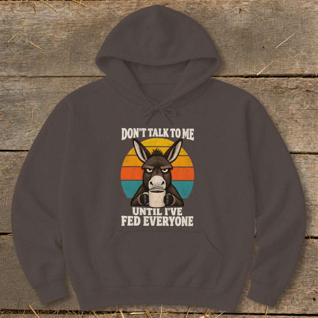 A dark brown hoodie featuring a grumpy donkey holding a coffee mug in front of a retro sunset graphic with the text, DON'T TALK TO ME UNTIL I'VE FED EVERYONE.
