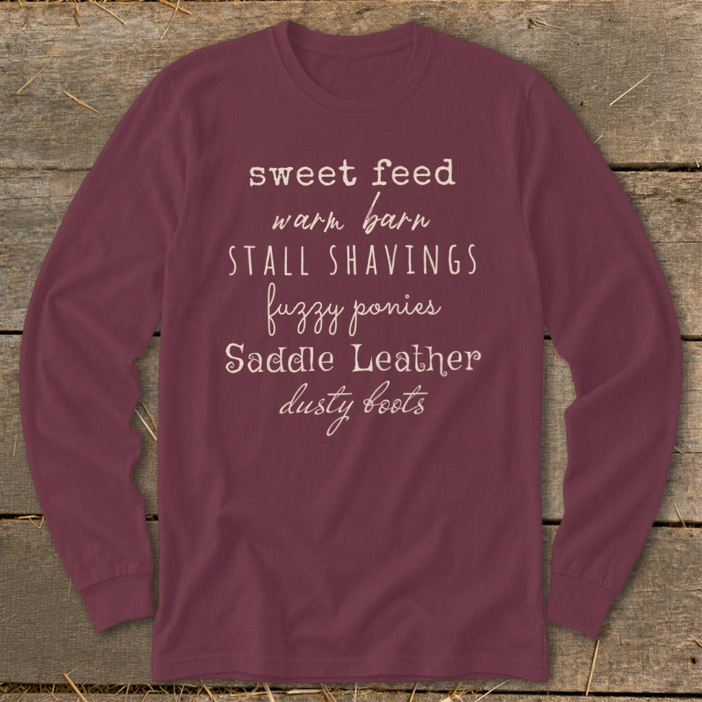 Burgundy long-sleeve shirt with white text listing horse-related terms: sweet feed, warm barn, stall shavings, fuzzy ponies, saddle leather, dusty boots.