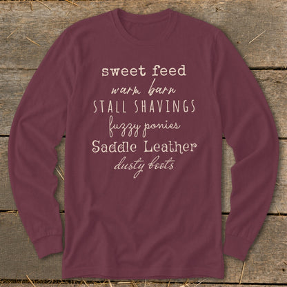 Burgundy long-sleeve shirt with white text listing horse-related terms: sweet feed, warm barn, stall shavings, fuzzy ponies, saddle leather, dusty boots.