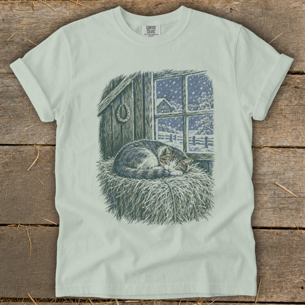 T-shirt graphic of a sleeping cat on hay in front of a barn window with snow falling.