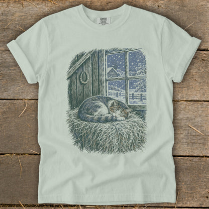 T-shirt graphic of a sleeping cat on hay in front of a barn window with snow falling.