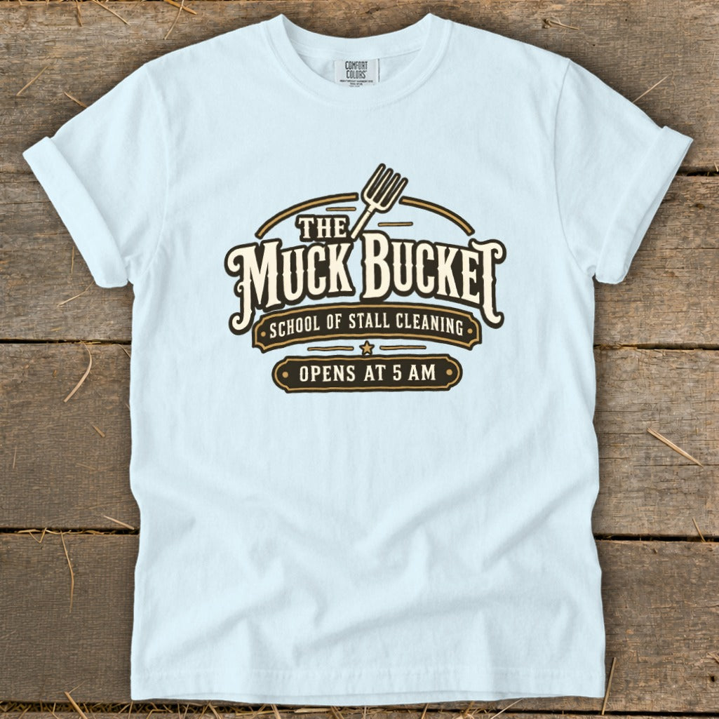 Light blue t-shirt with "The Muck Bucket School of Stall Cleaning Opens at 5 AM" logo.