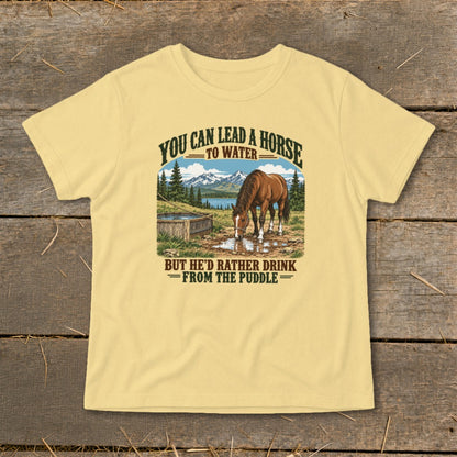 Light yellow t-shirt featuring a graphic of a horse drinking from a puddle with mountains and the text You Can Lead A Horse To Water But He'd Rather Drink From The Puddle.