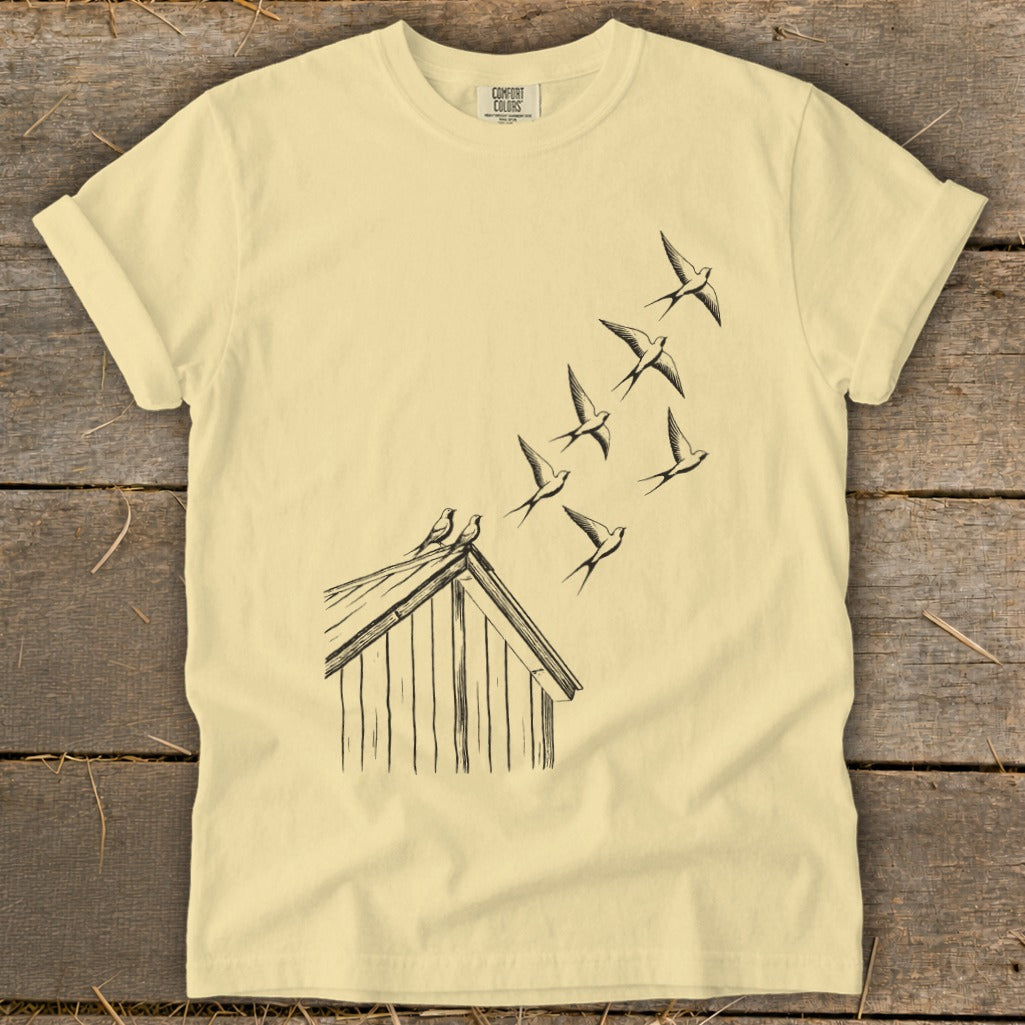 Light yellow t-shirt featuring a black line drawing of birds flying from a small wooden shed.