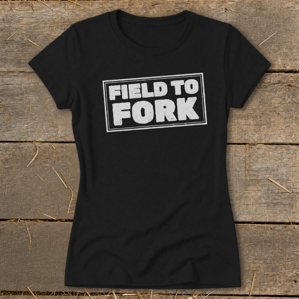 Black women's short-sleeve t-shirt with a white boxed graphic reading FIELD TO FORK laid flat on a rustic wooden surface.