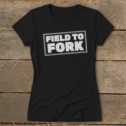 Black women's short-sleeve t-shirt with a white boxed graphic reading FIELD TO FORK laid flat on a rustic wooden surface.