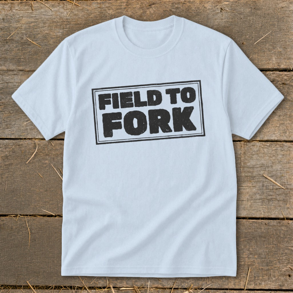 Light blue crewneck t-shirt featuring a black block-letter graphic reading FIELD TO FORK laid flat on a rustic wooden surface.