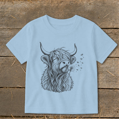 Highland cow blowing a dandelion on a light blue t-shirt.