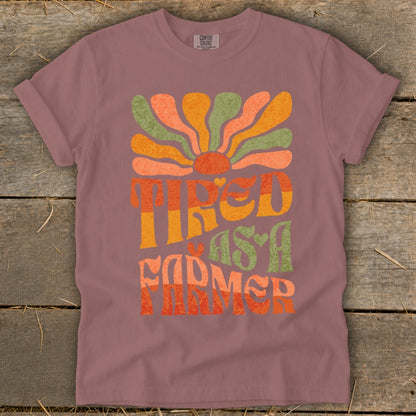 Tired as a Farmer text graphic with retro sunburst on muted pink t-shirt.