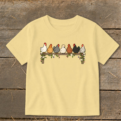 Light yellow children's t-shirt featuring an illustration of seven colorful chickens perched on a flower-covered fence.