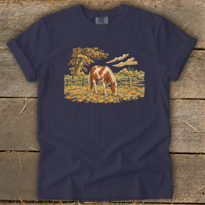 Navy t-shirt with a painting of a brown and white horse grazing in a field of fall leaves.