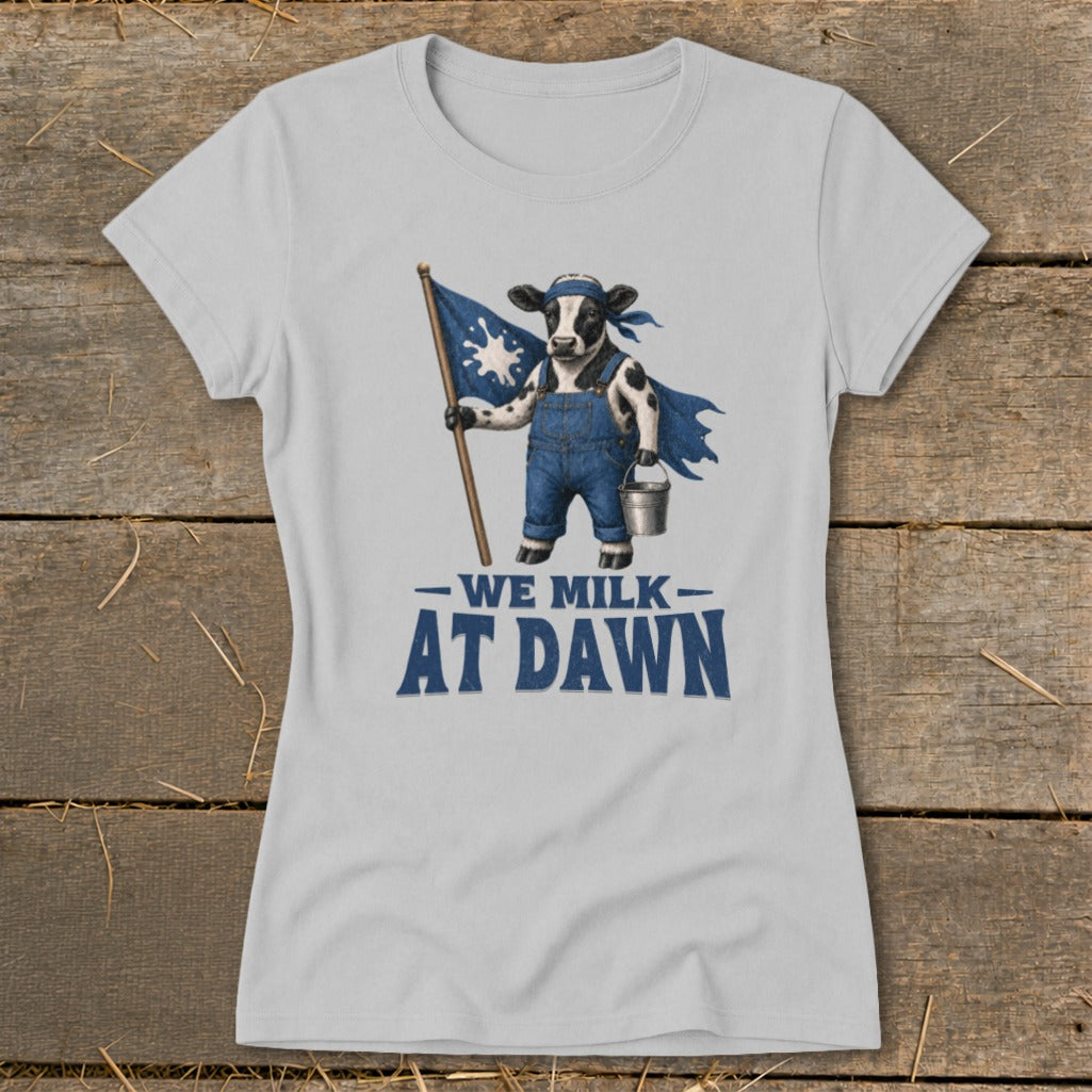 Light grey t-shirt with a cow wearing overalls and a cape, holding a flag and pail. Text reads "We Milk At Dawn."