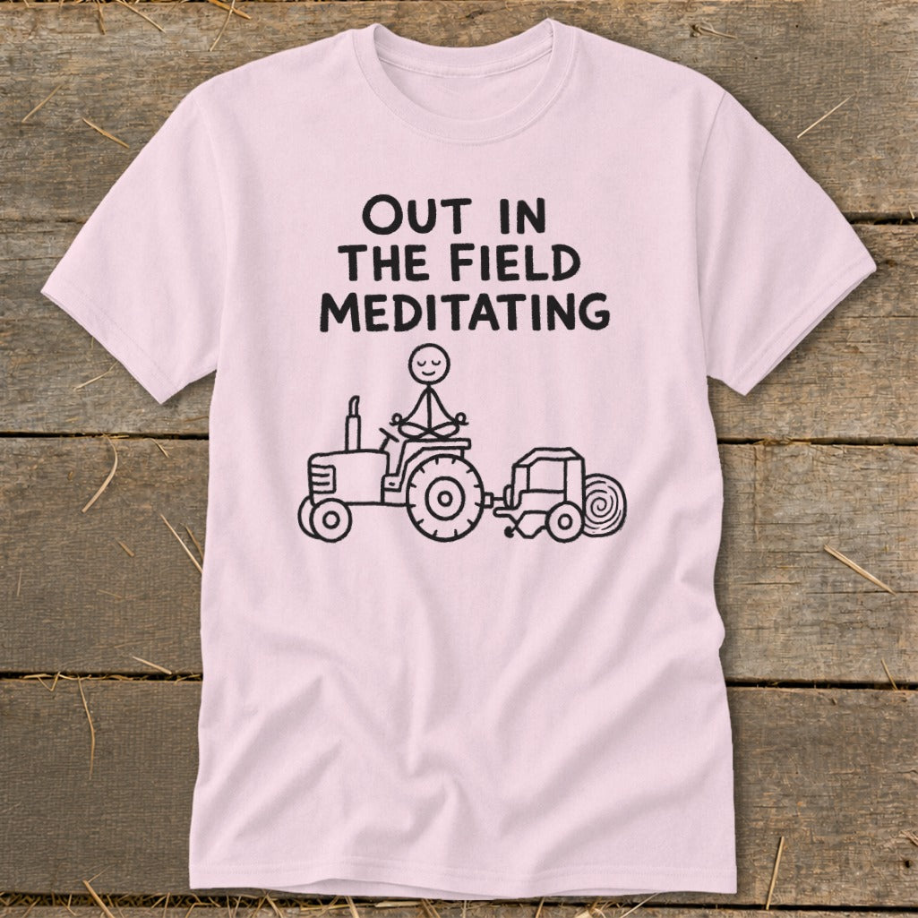 Pink t-shirt with black text "OUT IN THE FIELD MEDITATING" and stick figure on tractor.