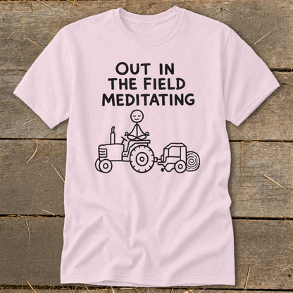 Pink t-shirt with black text "OUT IN THE FIELD MEDITATING" and stick figure on tractor.