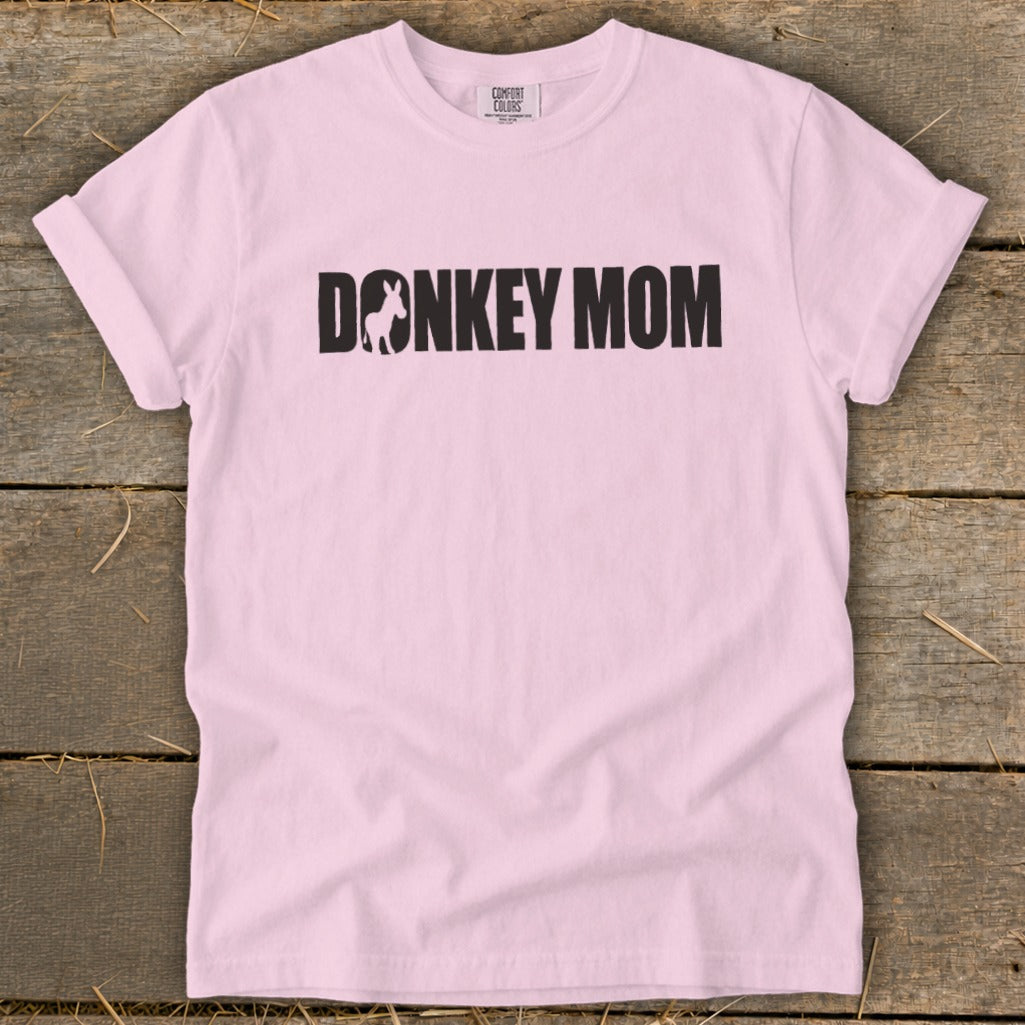 A light pink crewneck t-shirt with rolled sleeves lies flat on a rustic wooden background, featuring black text that reads DONKEY MOM with a small donkey silhouette inside the letter O.