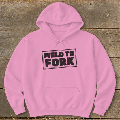 Light pink hooded sweatshirt with a front pocket and black text reading FIELD TO FORK centered on the chest, laid flat on a rustic wooden background.