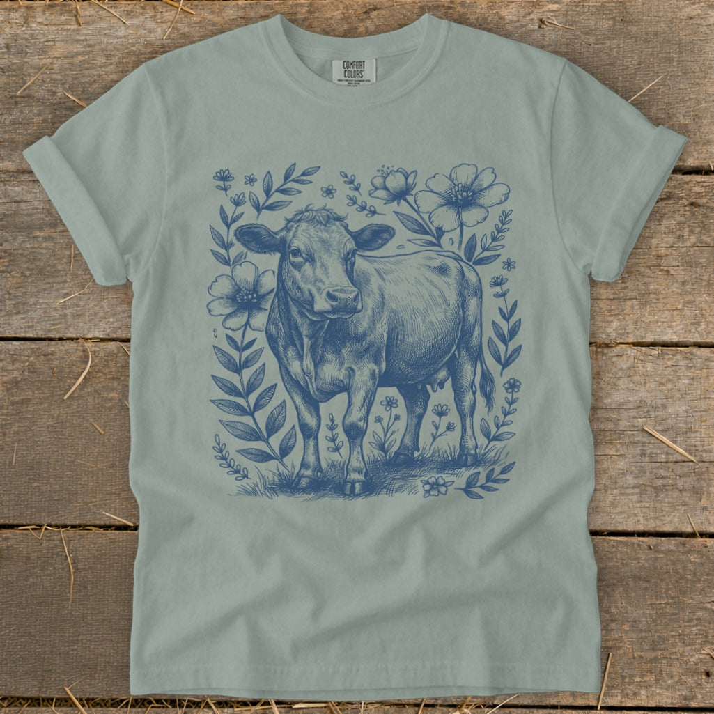 Blue line drawing of a cow surrounded by flowers on a pale green t-shirt.