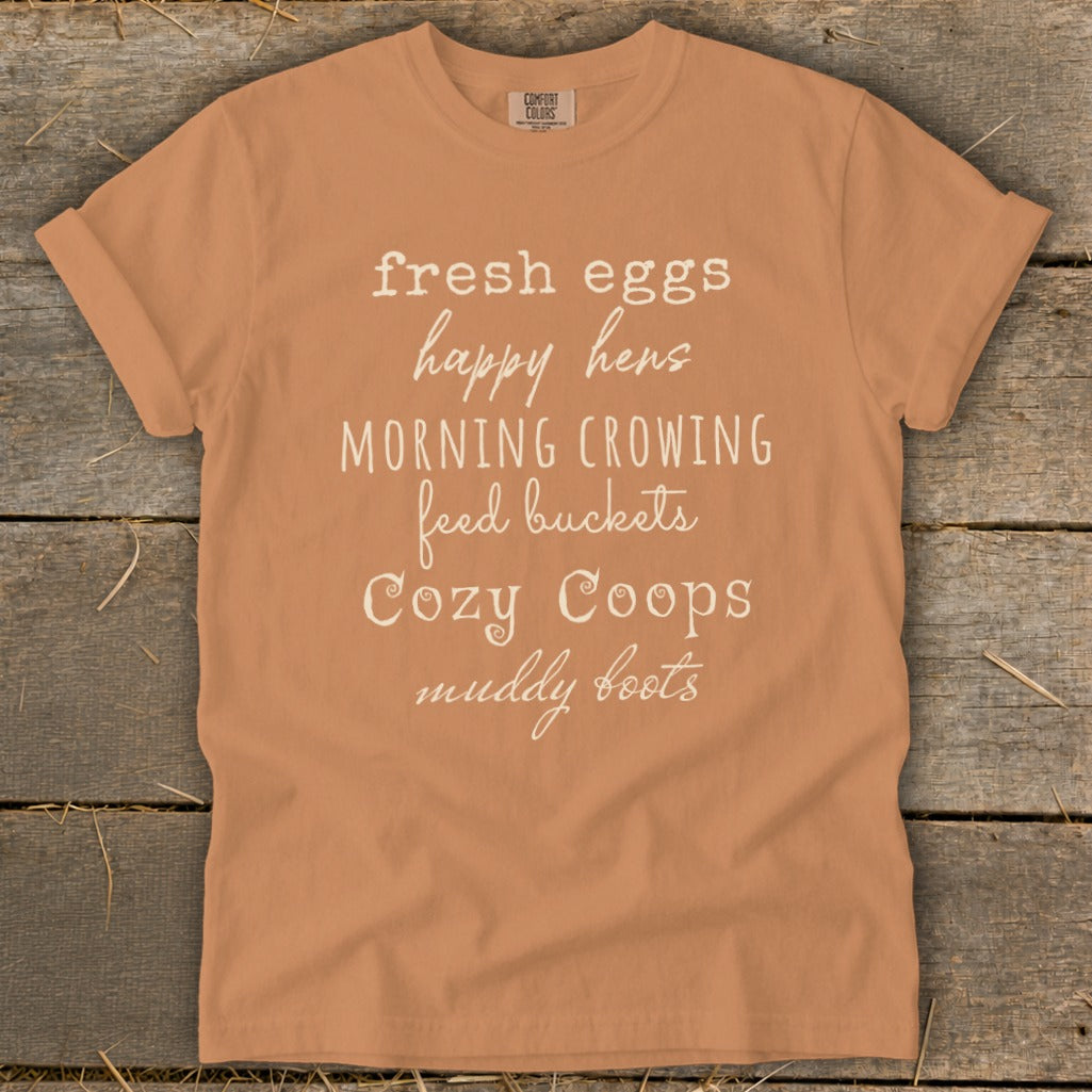 Peach t-shirt with white text: fresh eggs, happy hens, morning crowing, feed buckets, cozy coops, muddy boots.