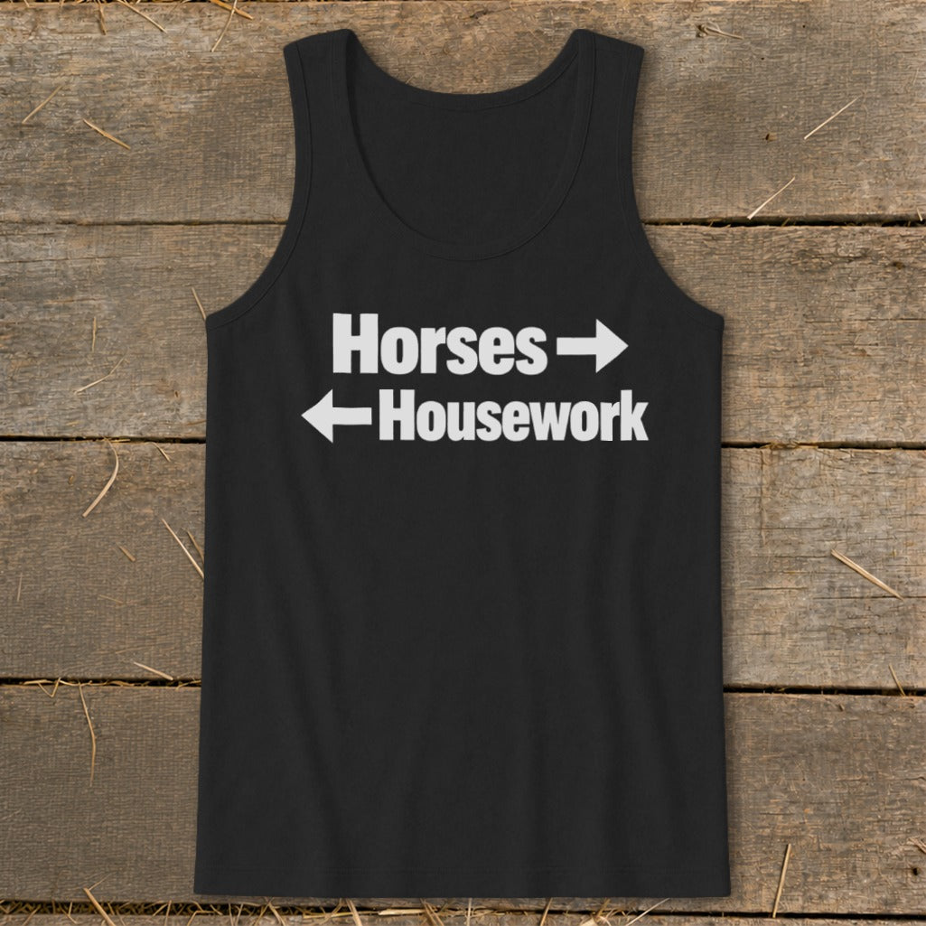 A black tank top with white text that reads Horses with a right arrow above Housework with a left arrow, laid flat on a rustic wooden background.
