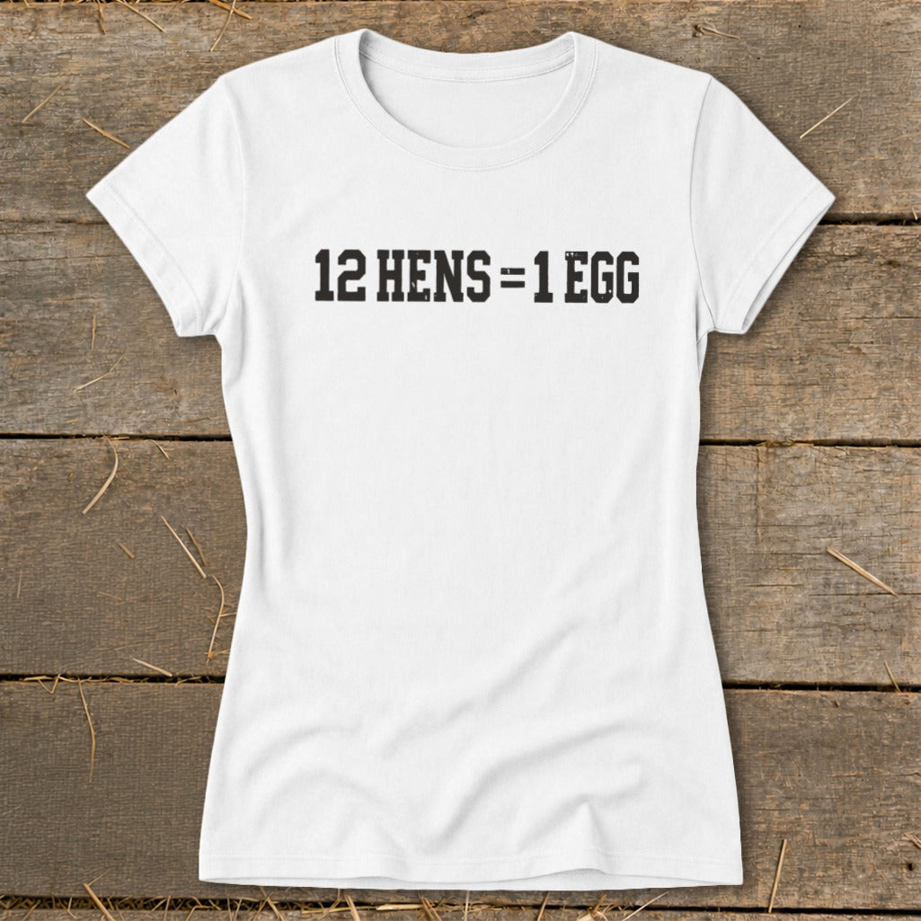 White short-sleeved women's t-shirt laid flat on a rustic wooden surface with black text reading 12 HENS = 1 EGG.