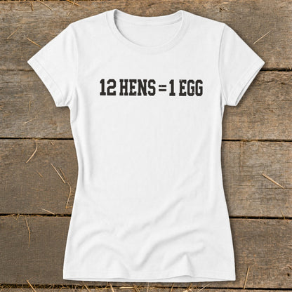 White short-sleeved women's t-shirt laid flat on a rustic wooden surface with black text reading 12 HENS = 1 EGG.