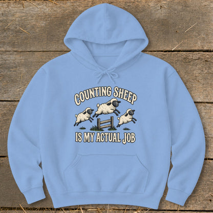 Light blue hoodie with cartoon sheep jumping over a fence and text "COUNTING SHEEP IS MY ACTUAL JOB".