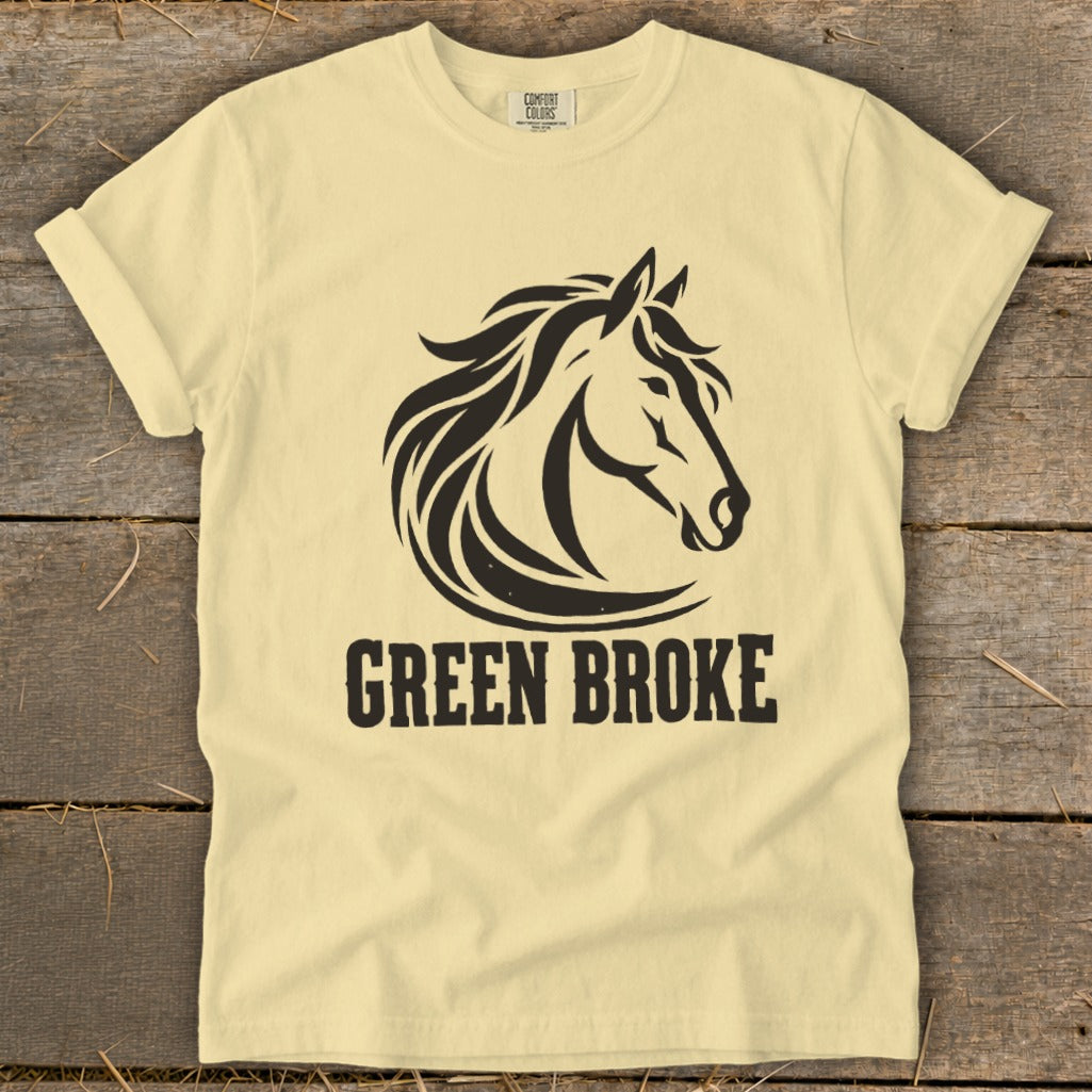 A cream-colored t-shirt with a black horse head graphic and the text "GREEN BROKE" lying on a wooden background.