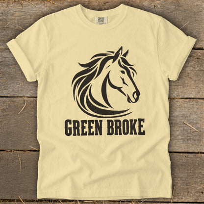 A cream-colored t-shirt with a black horse head graphic and the text "GREEN BROKE" lying on a wooden background.
