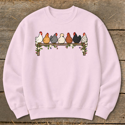 Light pink crewneck sweatshirt featuring a graphic of seven colorful cartoon chickens sitting on a flower-adorned fence.