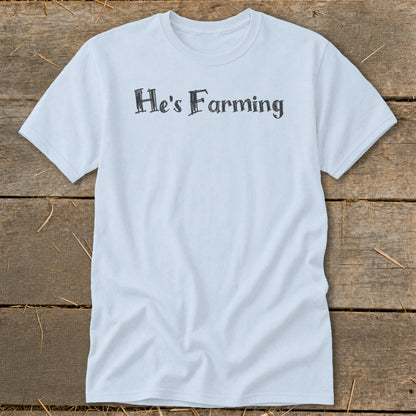 Light blue t-shirt with "He's Farming" text in charcoal gray, laid on rustic wooden planks.