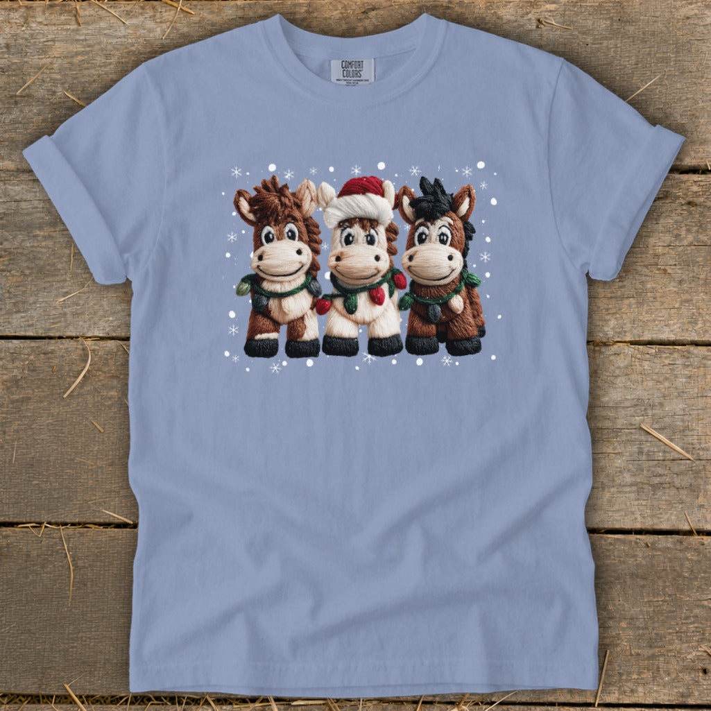 Three cartoon horses in Christmas attire with snowflakes on a light blue t-shirt.