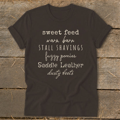 Dark t-shirt with white text listing "sweet feed, warm barn, stall shavings, fuzzy ponies, Saddle Leather, dusty boots."