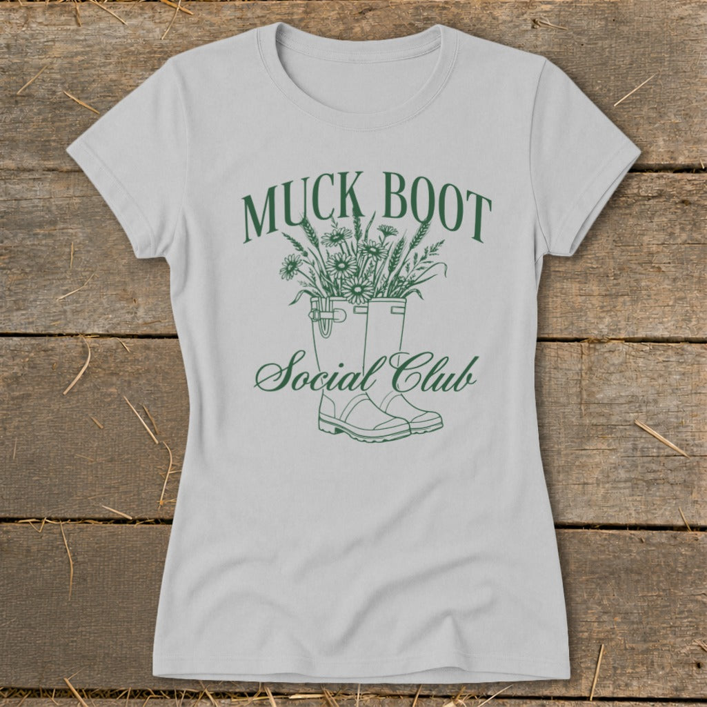Light grey t-shirt with green graphic of muck boots filled with flowers and wheat.