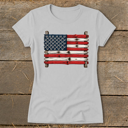 A light gray women's t-shirt features a graphic of the American flag made of painted wooden planks with horseshoes.