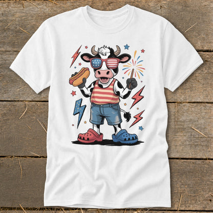 Cartoon cow wearing American flag sunglasses, holding hot dog and sparkler on white t-shirt.
