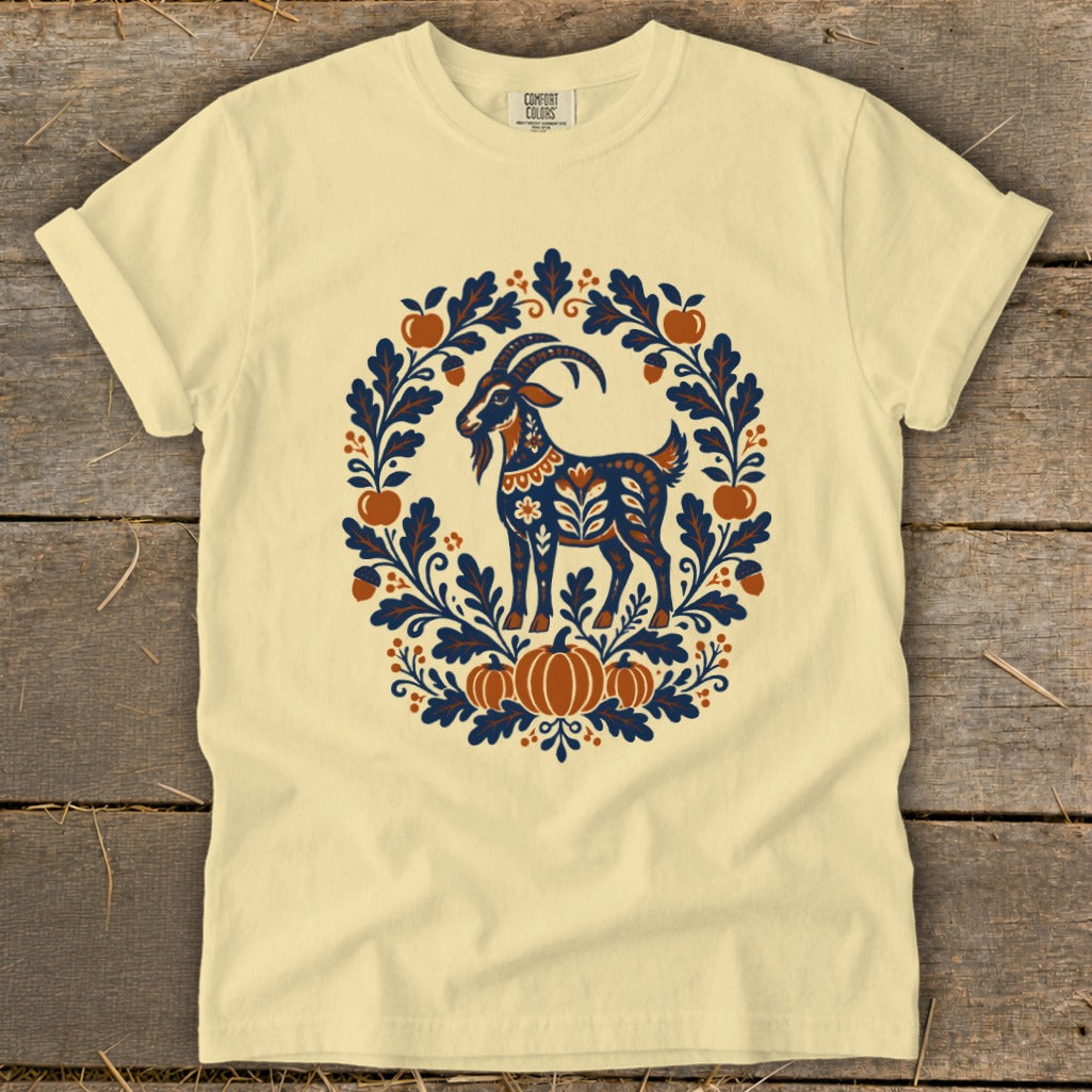 A cream-colored t-shirt with a blue and orange folk art illustration of a goat surrounded by leaves and pumpkins.