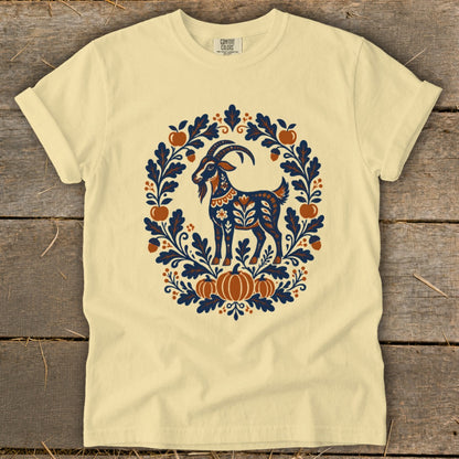 A cream-colored t-shirt with a blue and orange folk art illustration of a goat surrounded by leaves and pumpkins.