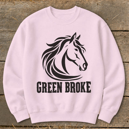 A light pink crewneck sweatshirt featuring a black graphic of a horse head and the text GREEN BROKE on a wooden surface.