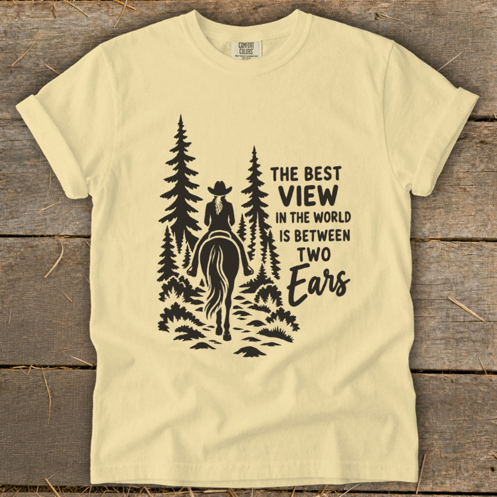 A light yellow t-shirt with a graphic of a horseback rider in pine trees and the text THE BEST VIEW IN THE WORLD IS BETWEEN TWO EARS.